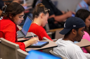 Students take notes in a large lecture hall, facing away from the camera
